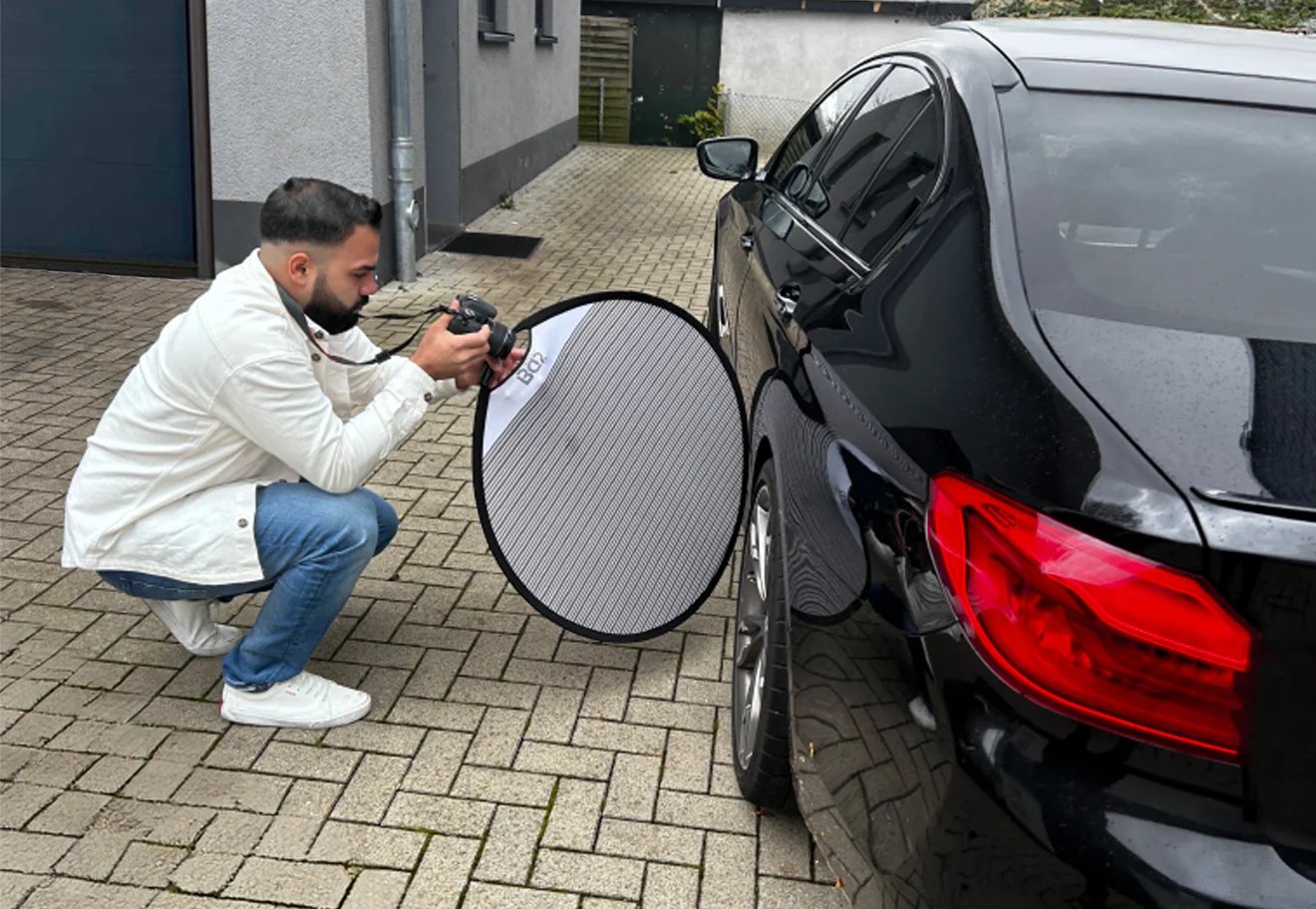 Man holding a reflector disc while photographing the side of a black car in a driveway